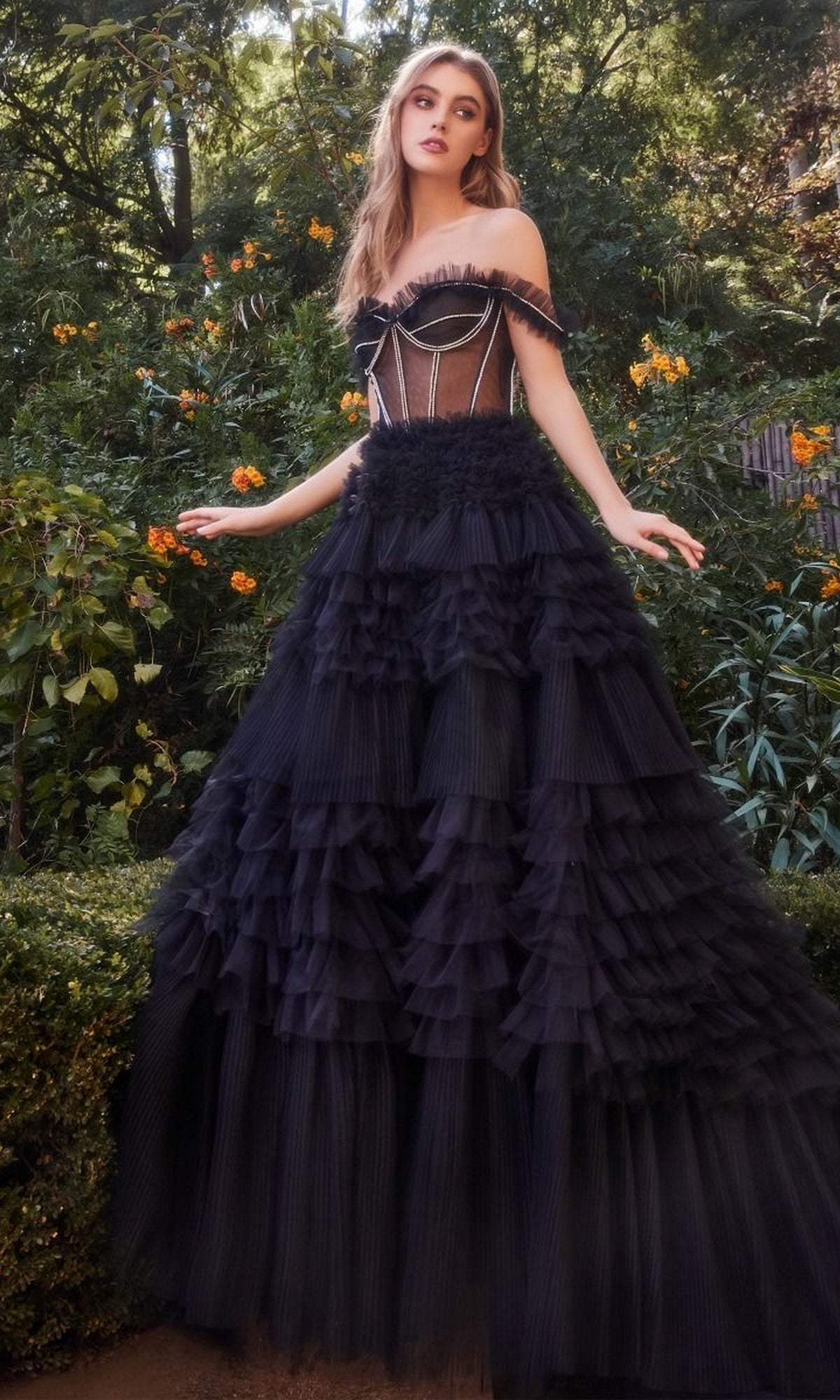 A woman stands outdoors in a garden, wearing the Pastel Off-the-Shoulder Ruffled Ball Gown A1150 featuring a fitted sheer bodice and layered tulle skirt.
